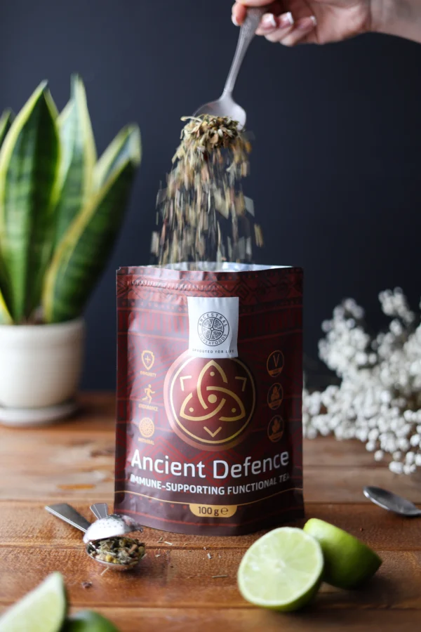 Kräutertee-Mischung Ancient Defence fällt vom Löffel zurück in die Verpackung – sichtbar naturbelassen mit Limette und Teesieb auf Holztisch.