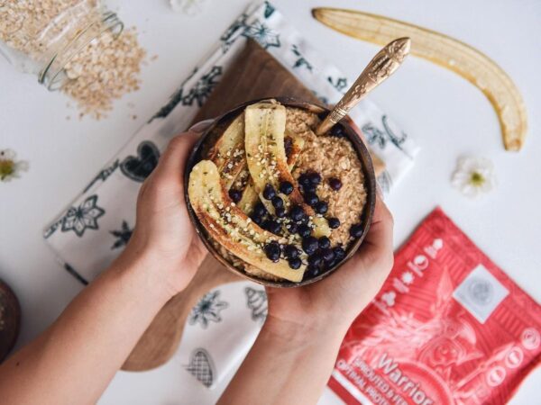Haferbrei mit proteinreichem Warrior-Pulver in einer Schüssel, getoppt mit gegrillter Banane und Heidelbeeren, gehalten von zwei Händen
